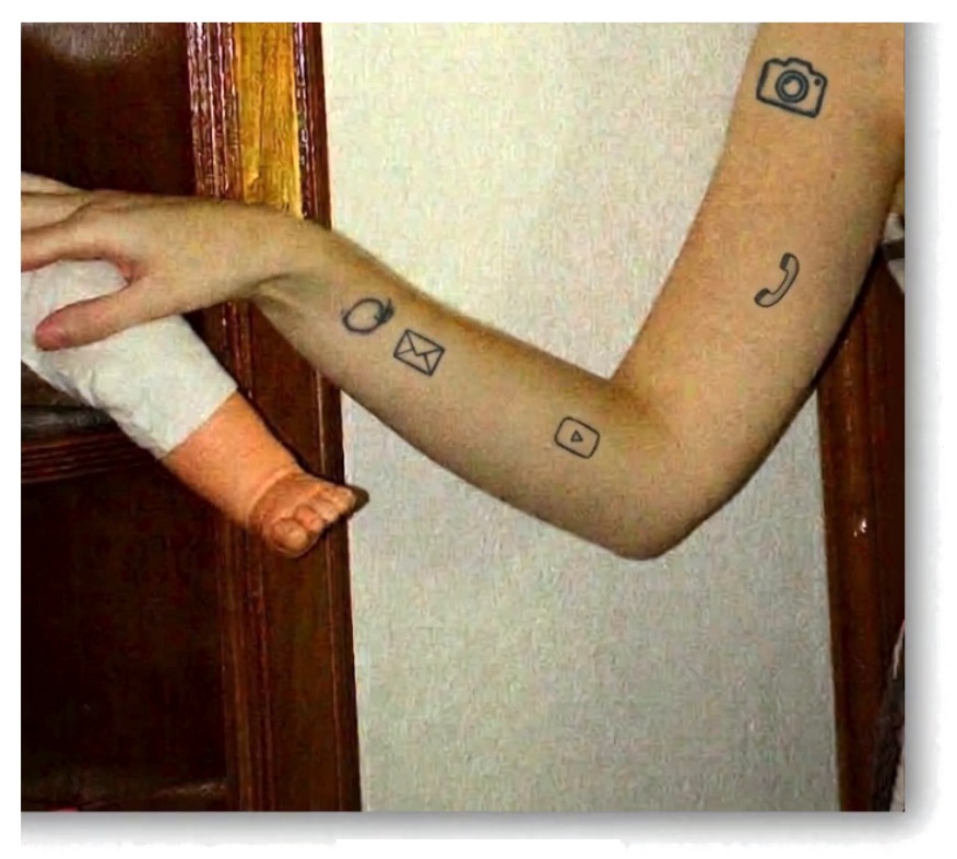 An arm holding a doll's hand, with small icons drawn on the skin like tattoos